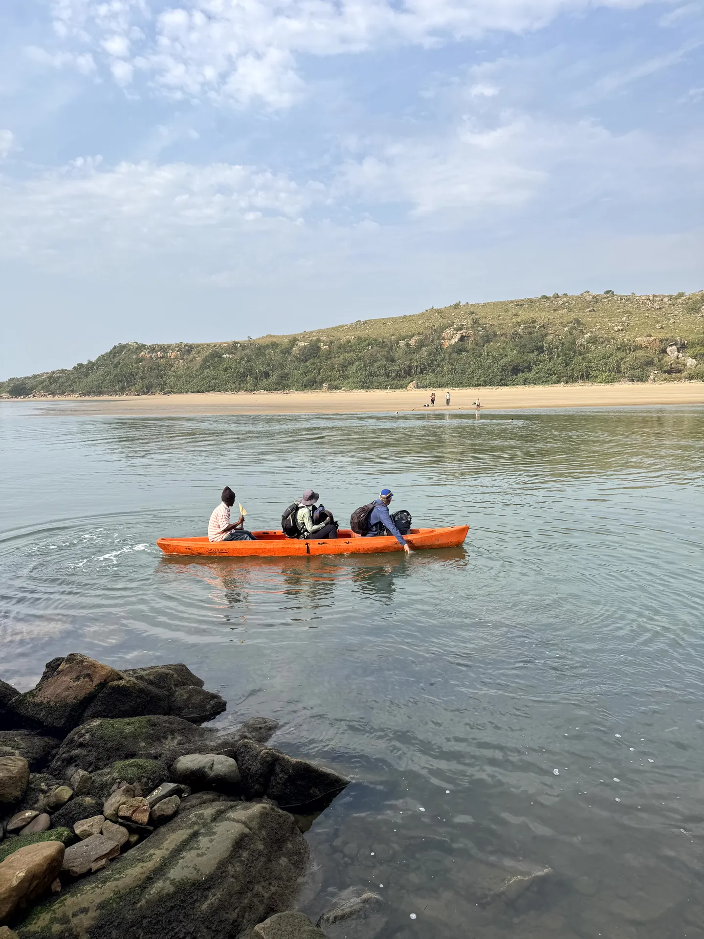 Orange kayak crossing calm estuary waters with sandy beach backdrop